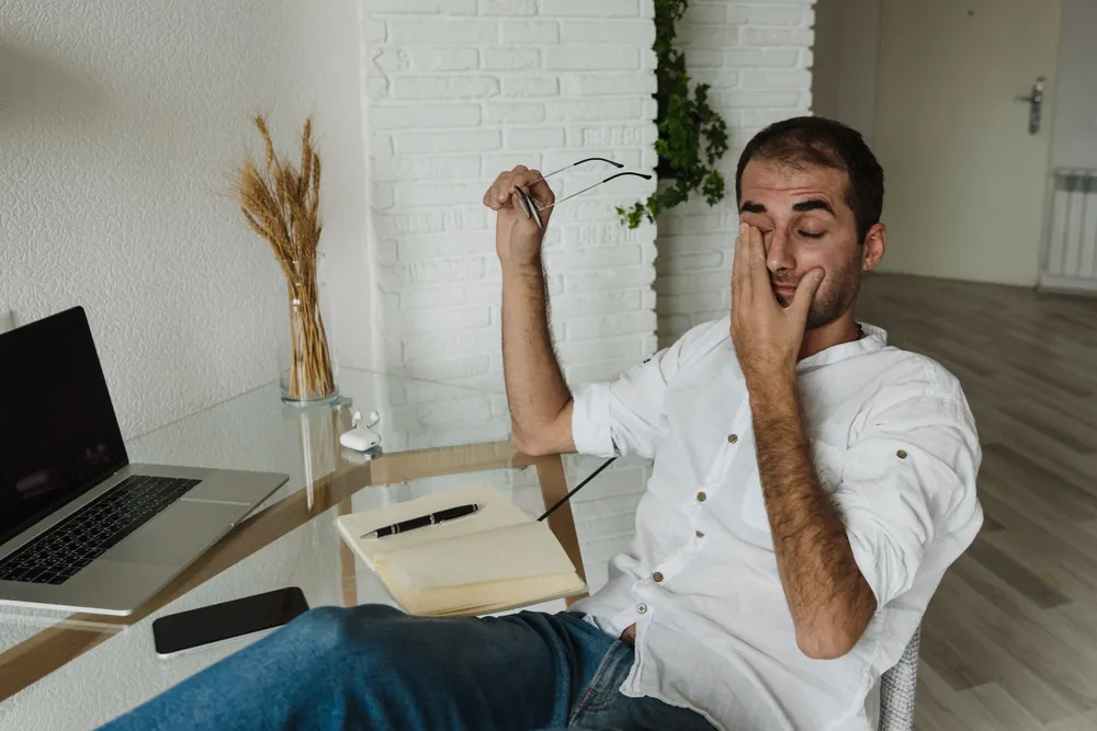 Dizüstü bilgisayarın başında gözlüğünü çıkarıp gözünü ovuşturan genç bir adam